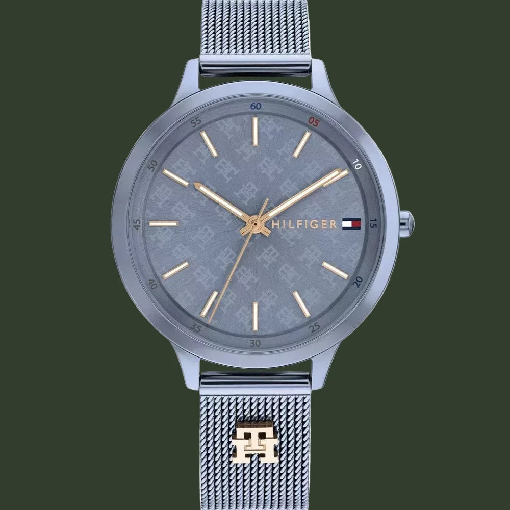 A Tommy Hilfiger men's analog watch with a silver-tone stainless steel case and mesh strap, featuring a textured gray dial with gold-tone hands and indices, and the brand's signature flag logo on the dial.