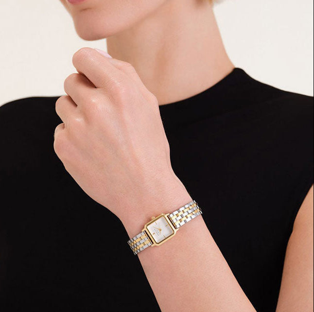 A close-up of a woman wearing a gold-tone watch with a silver-tone link bracelet and a white dial, shown against a black top and light background.