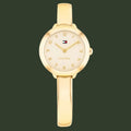 A gold-tone Tommy Hilfiger bangle watch with a round bezel, cream-colored dial, and a matching gold bangle strap, shown against a plain white background.