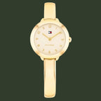 A gold-tone Tommy Hilfiger bangle watch with a round bezel, cream-colored dial, and a matching gold bangle strap, shown against a plain white background.